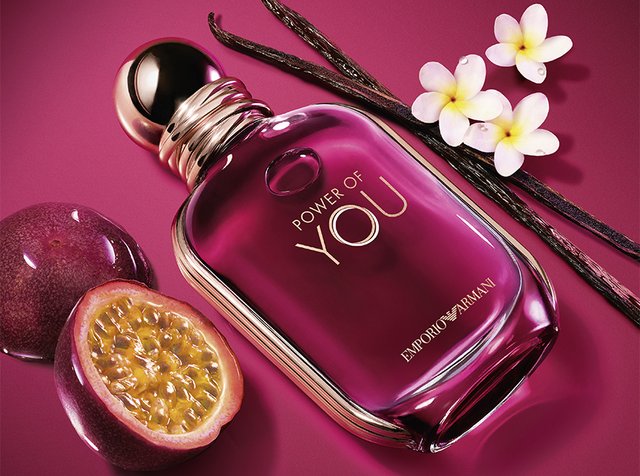 Bouteille de parfum Purple Emporio Armani Power of You à côté de fruit de la passion, de gousses de vanille et de fleurs de frangipanier blanches sur un fond violet.