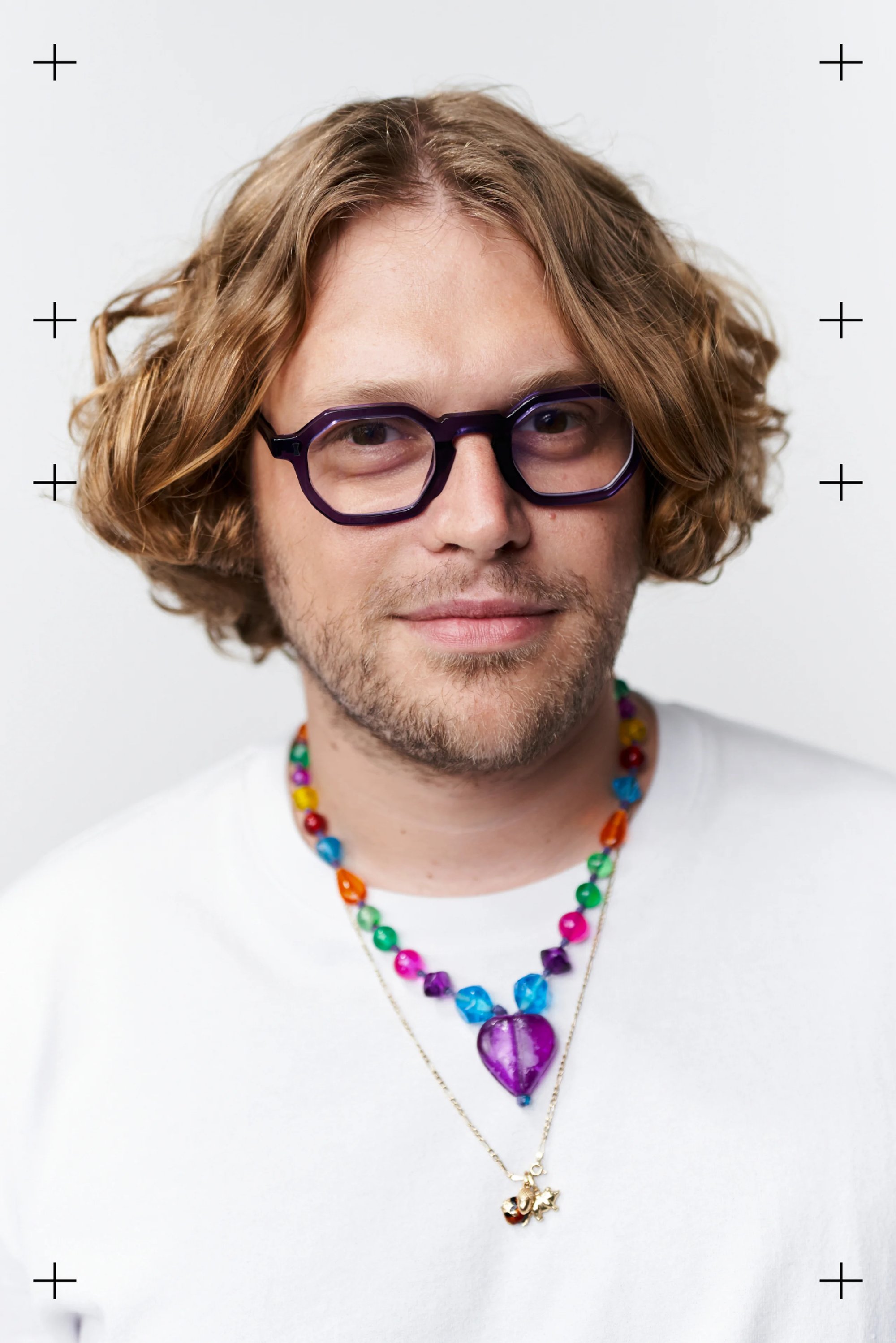 Una persona con cabello ondulado hasta los hombros y gafas sonríe, vistiendo una camisa blanca adornada con coloridos collares de cuentas.