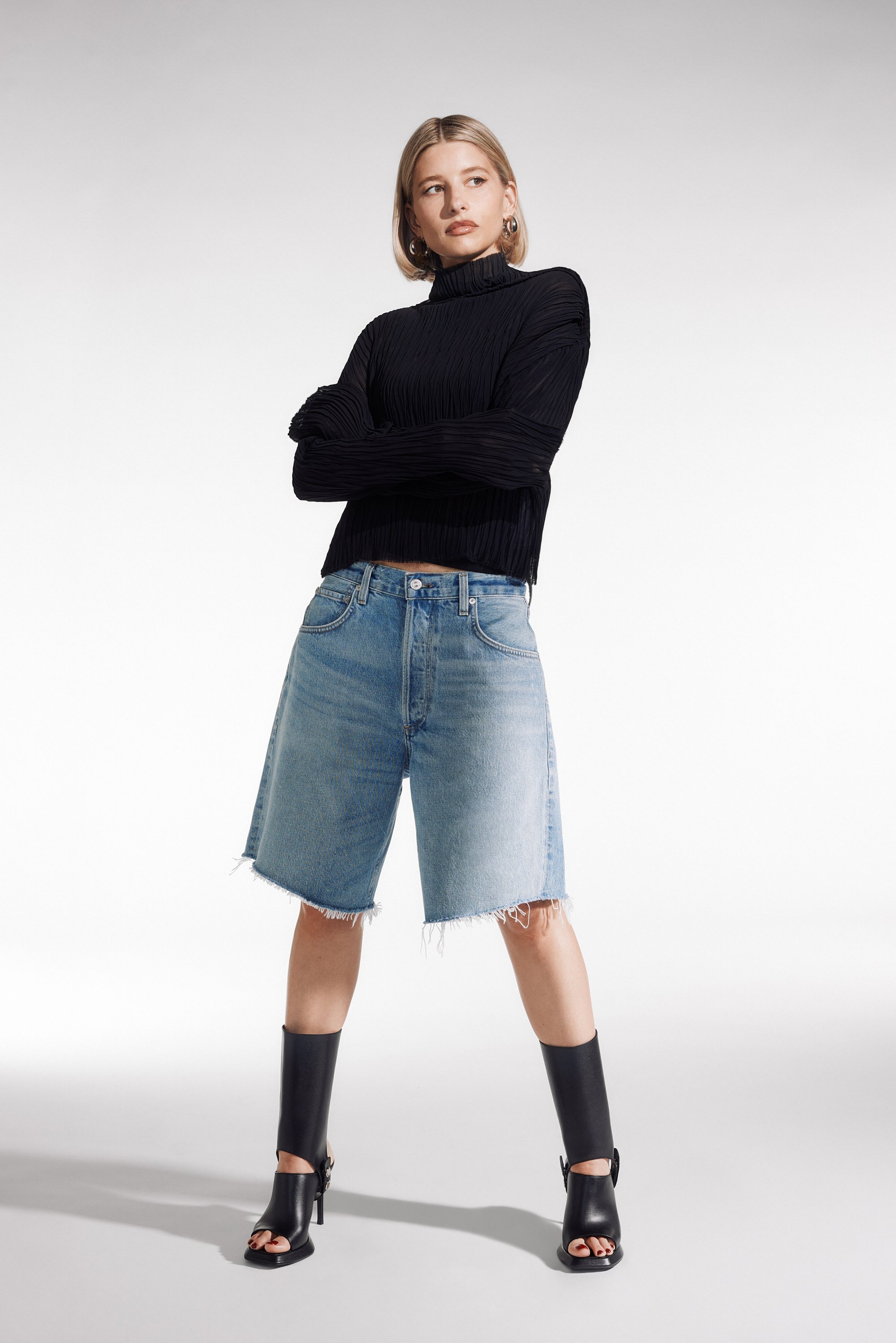 Woman posing confidently with arms crossed, wearing a black pleated top, blue denim shorts, and black high-heeled sandals.