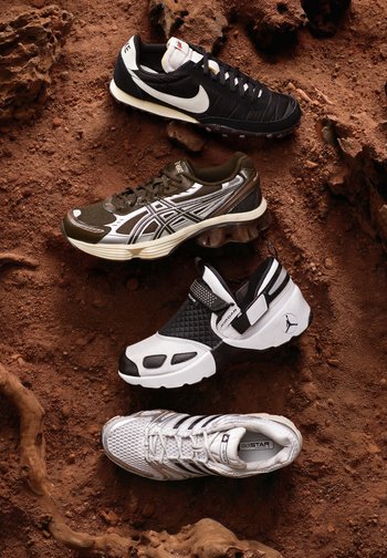 Cuatro zapatillas deportivas diferentes en negro, marrón, blanco y plateado dispuestas sobre un terreno rocoso rojizo con suelo texturizado y pequeñas piedras.