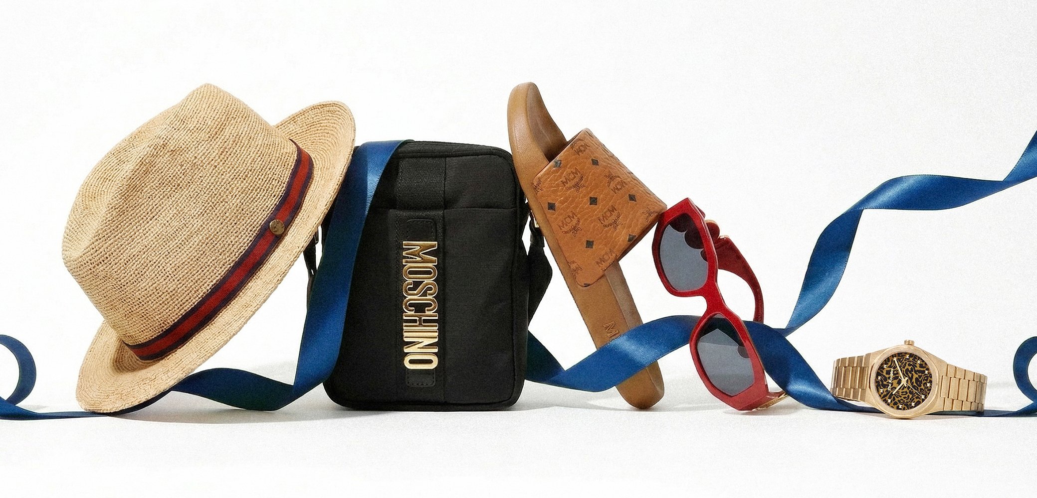 Straw hat, black Moschino bag, brown slide sandals, red sunglasses, and beige watch with blue ribbon on a white background.