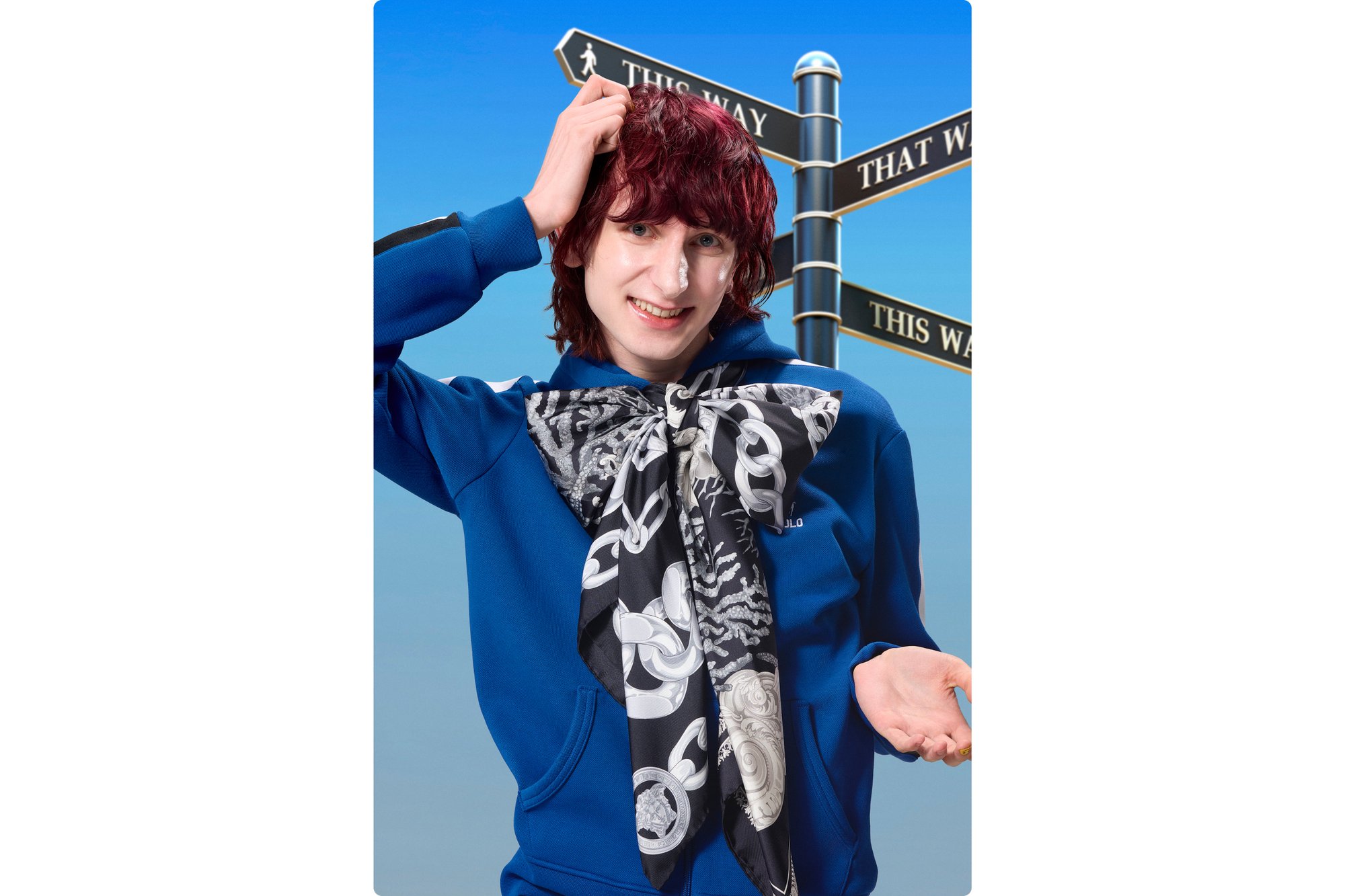 Una persona con i capelli rossi indossa un felpa blu e una sciarpa con motivo, posando davanti a segnali direzionali contro un cielo sereno.