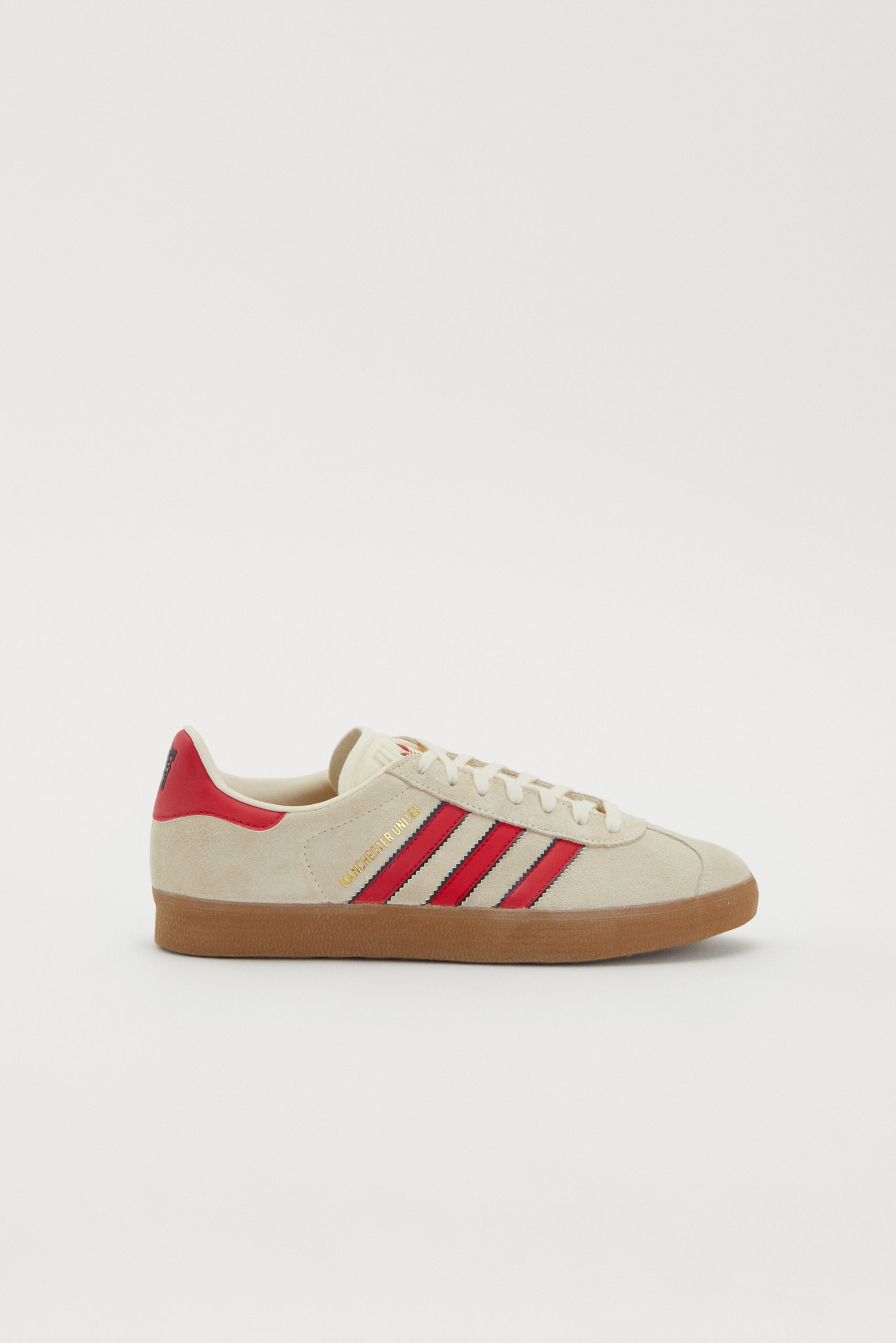Beige suède sneaker met rode en zwarte strepen, voorzien van een tan rubberen zool en gouden tekst die "Manchester" zegt. Zijaanzicht.