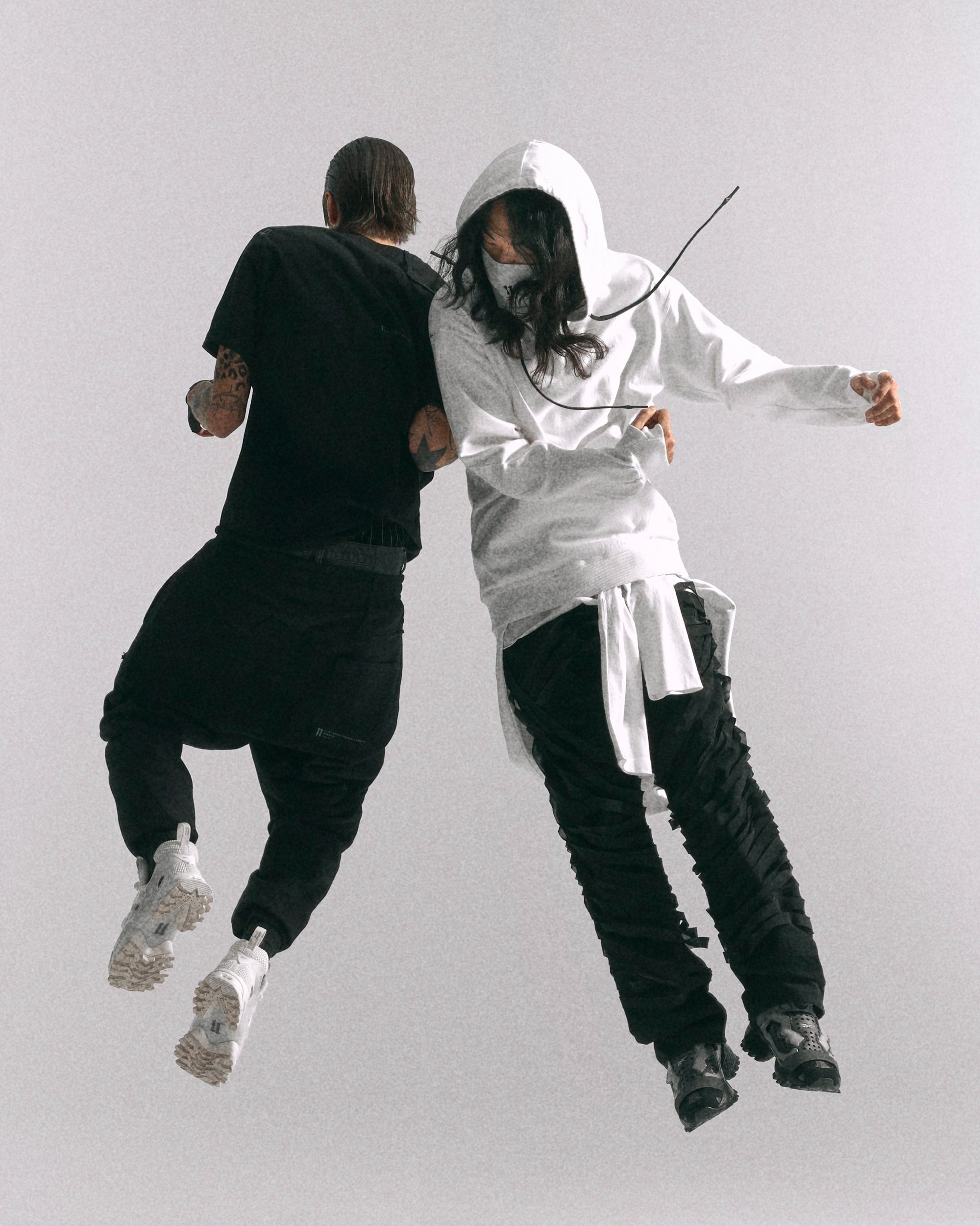 Dos personas saltan frente a un fondo liso, vestidas con atuendos contrastantes: una en negro y la otra en blanco, adoptando poses dinámicas.