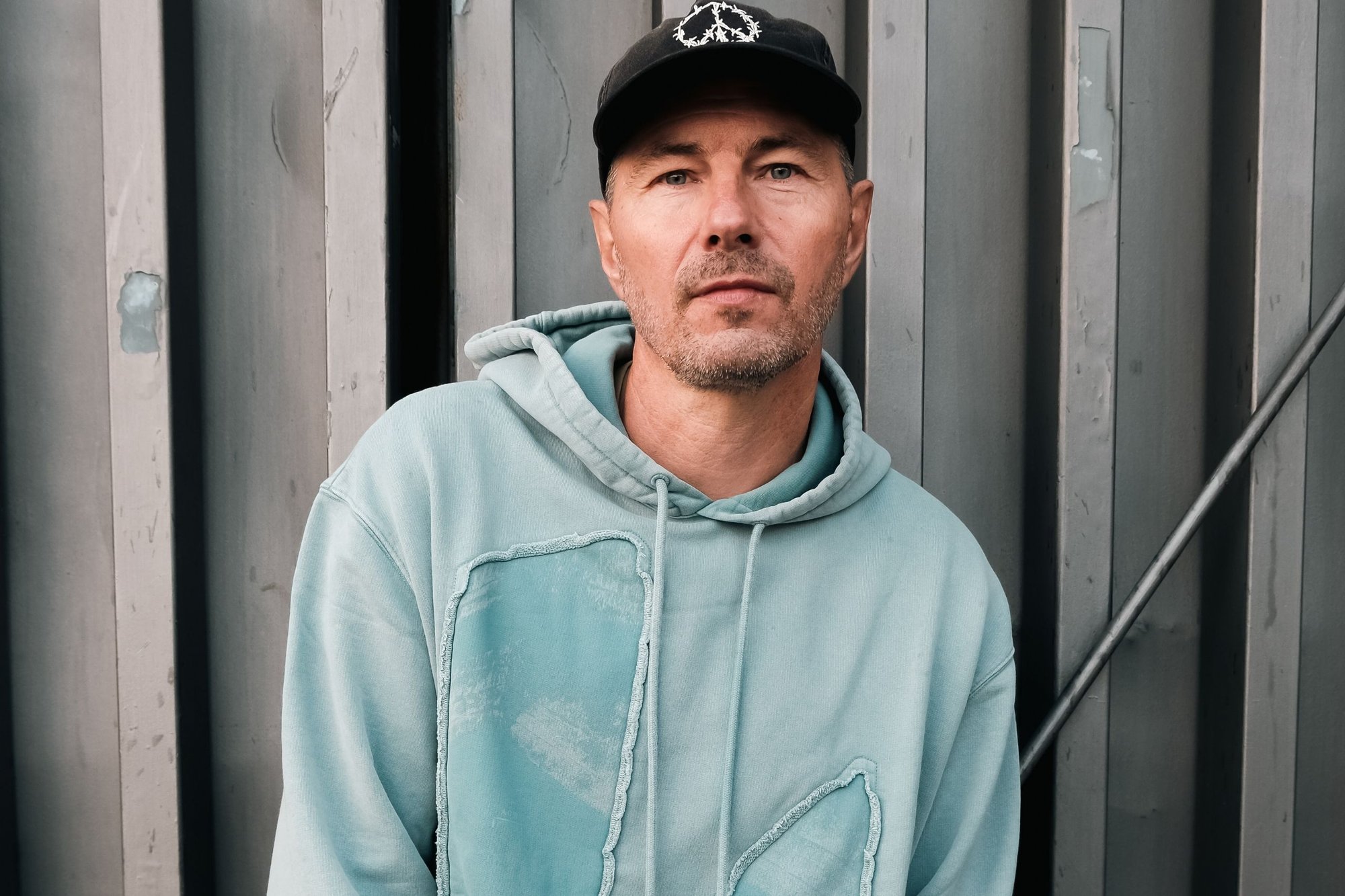 Mintgrön hoodie med en texturerad, abstrakt patchdesign, utrustad med en dragsko i huvan. Bärs med en svart keps mot en grå bakgrund.