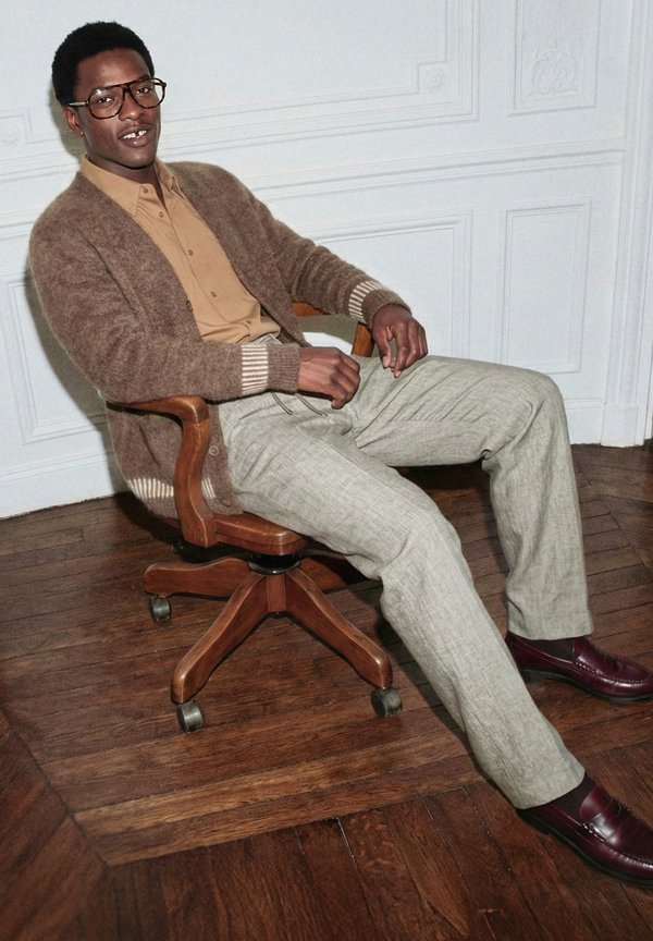 Άνδρας με γυαλιά, καφέ ζακέτα, μπεζ πουκάμισο και ανοιχτό γκρι παντελόνι, καθισμένος σε ξύλινη περιστρεφόμενη καρέκλα σε σκούρο ξύλινο πάτωμα.