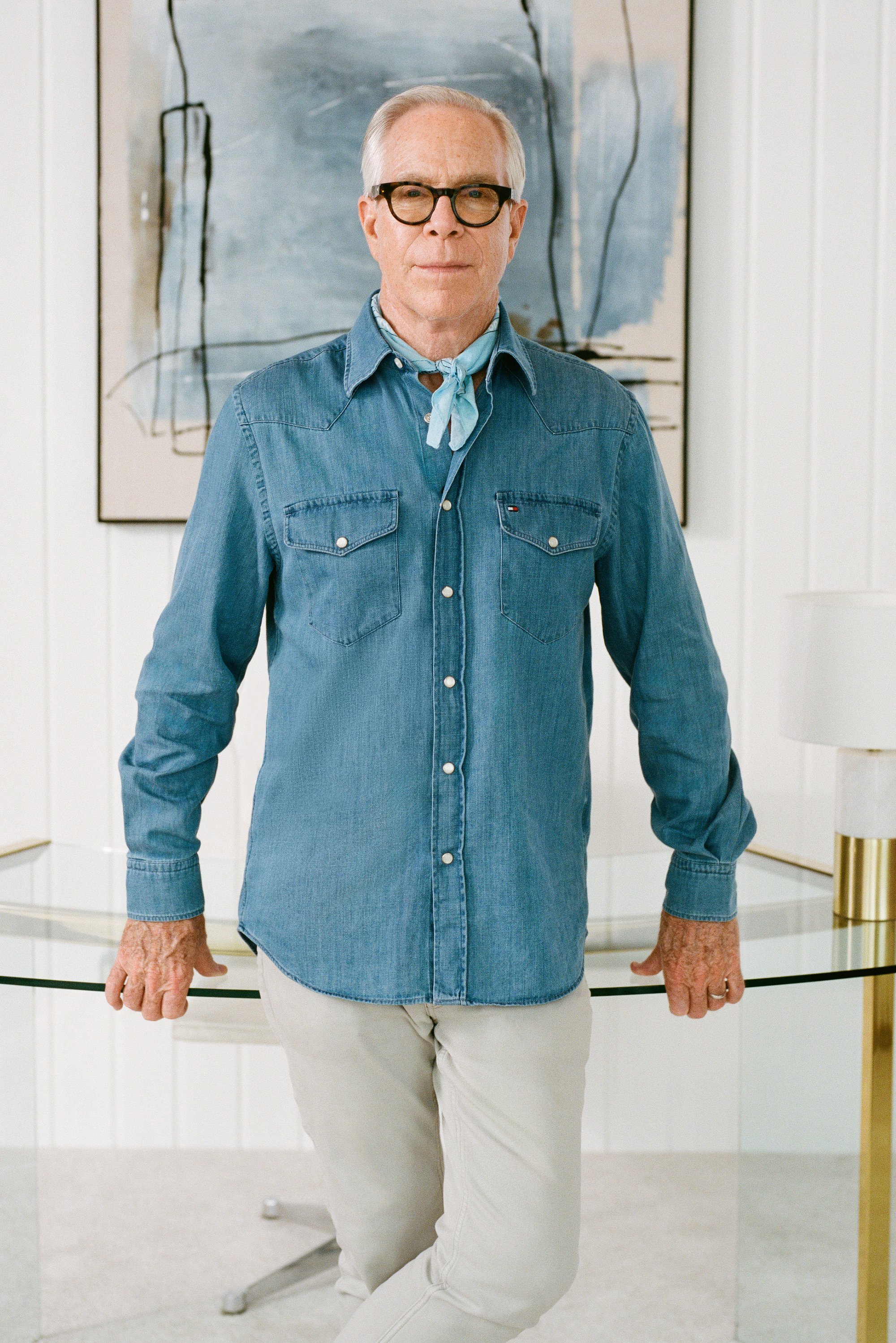 Denim knoopshirt met twee borstzakken, lichtblauwe kleur, gedragen met een lichtblauwe sjaal om de nek, gecombineerd met beige broek, staande bij een glazen tafel.