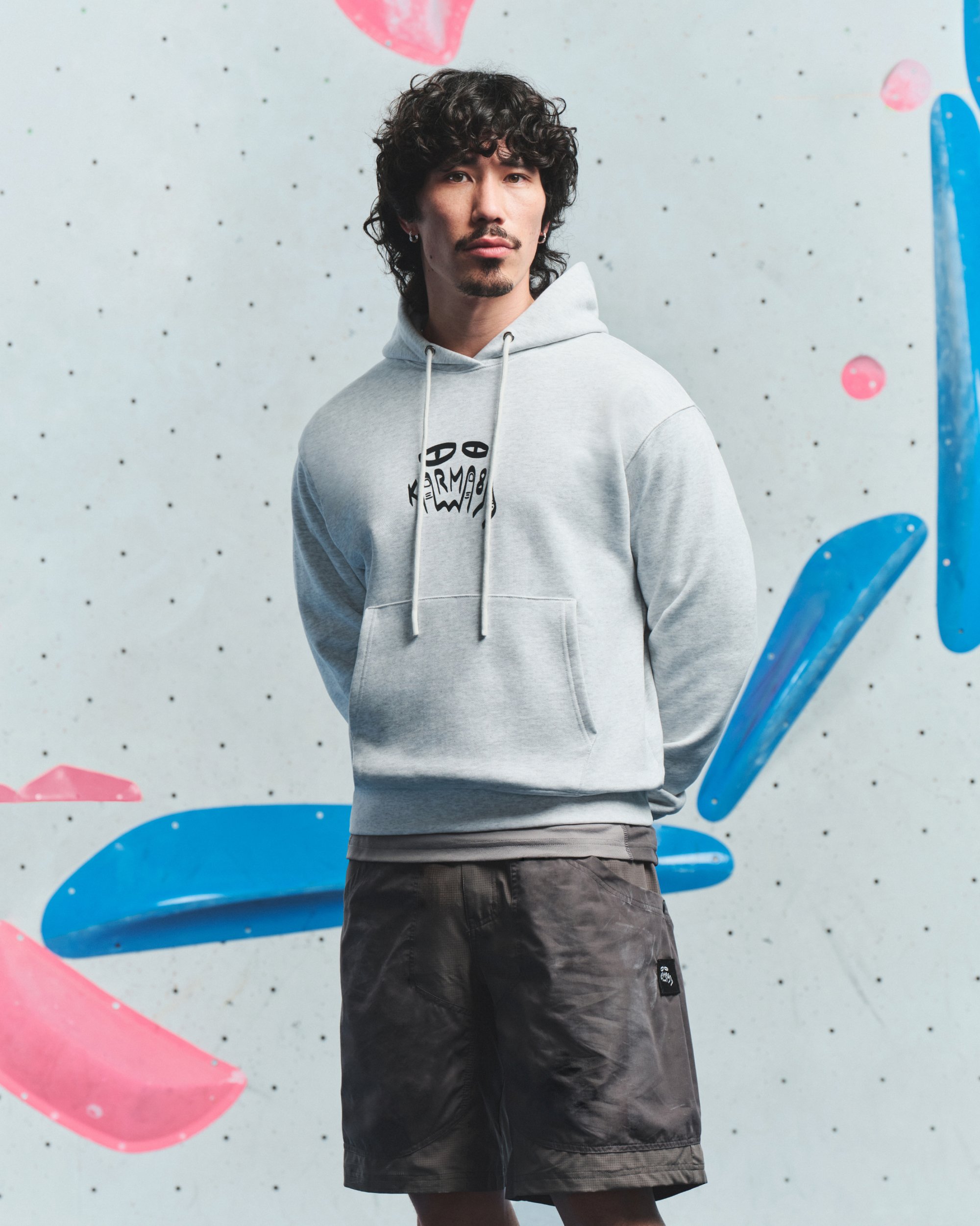 Hombre con cabello rizado y perilla, vistiendo una sudadera con capucha gris claro y pantalones cortos oscuros, se encuentra de pie con las manos detrás de la espalda frente a una pared de escalada con agarres azules y rosas.