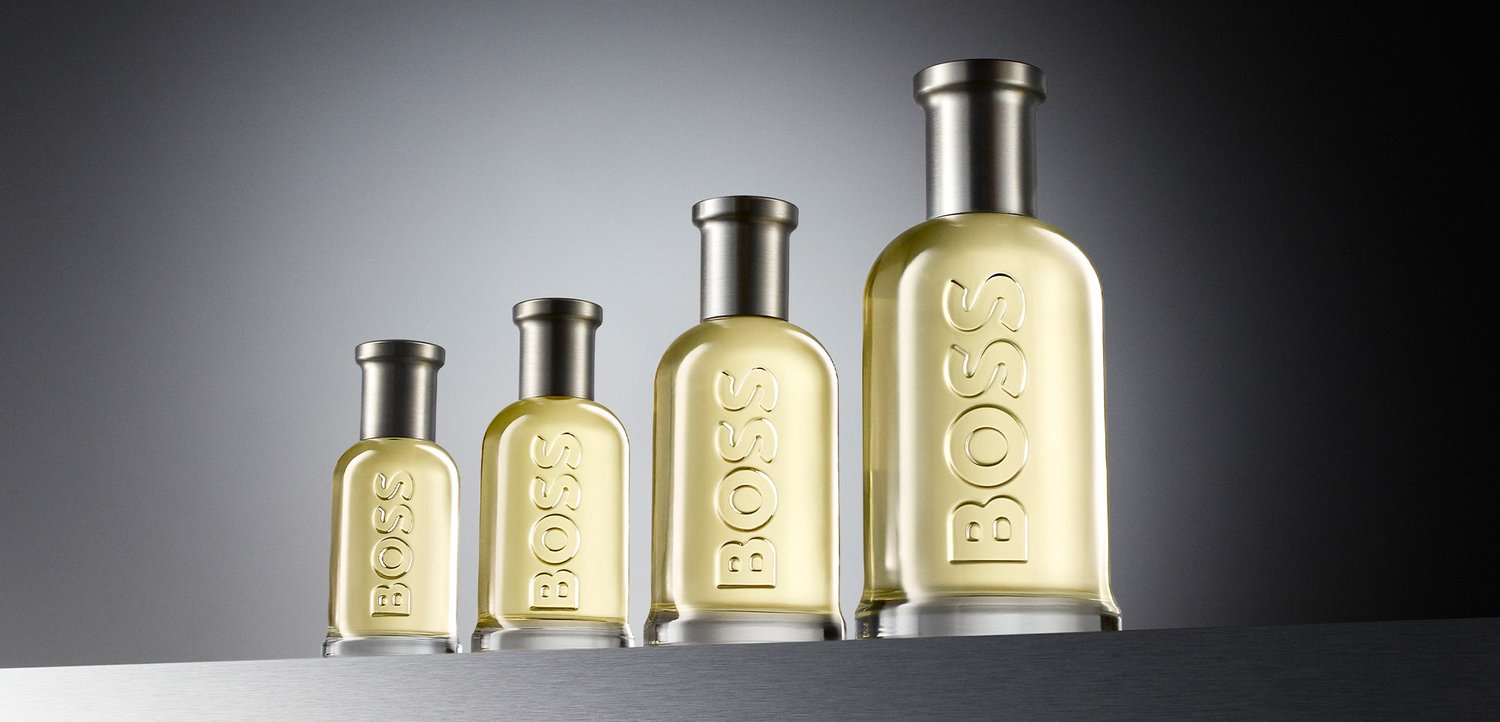 Quattro flaconi di profumo in vetro trasparente con tappi argentati e liquido giallo all'interno, ciascuno etichettato "BOSS,", disposti in ordine crescente da sinistra a destra.