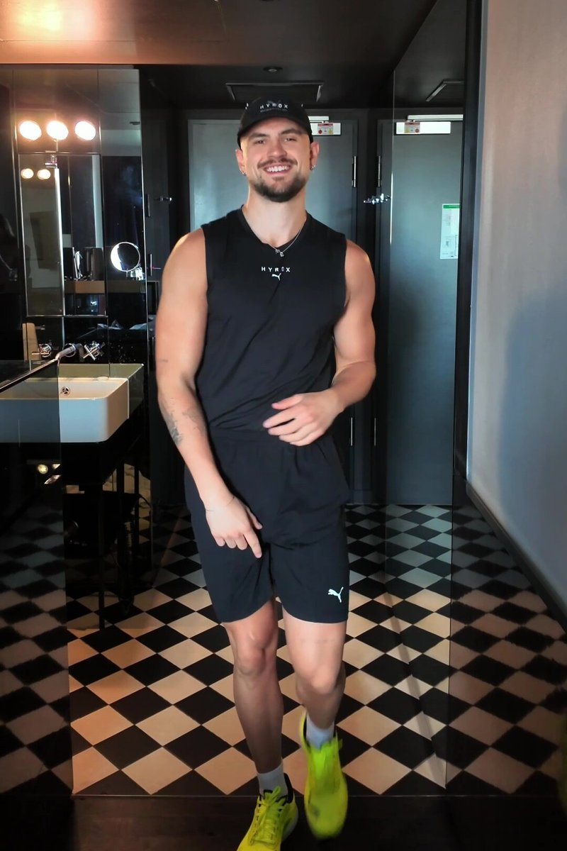 Černý tílko bez rukávů a šortky s bílým logem, doplněné jasně žlutými sportovními botami. Černobílá kostkovaná podlaha; prostředí koupelny.