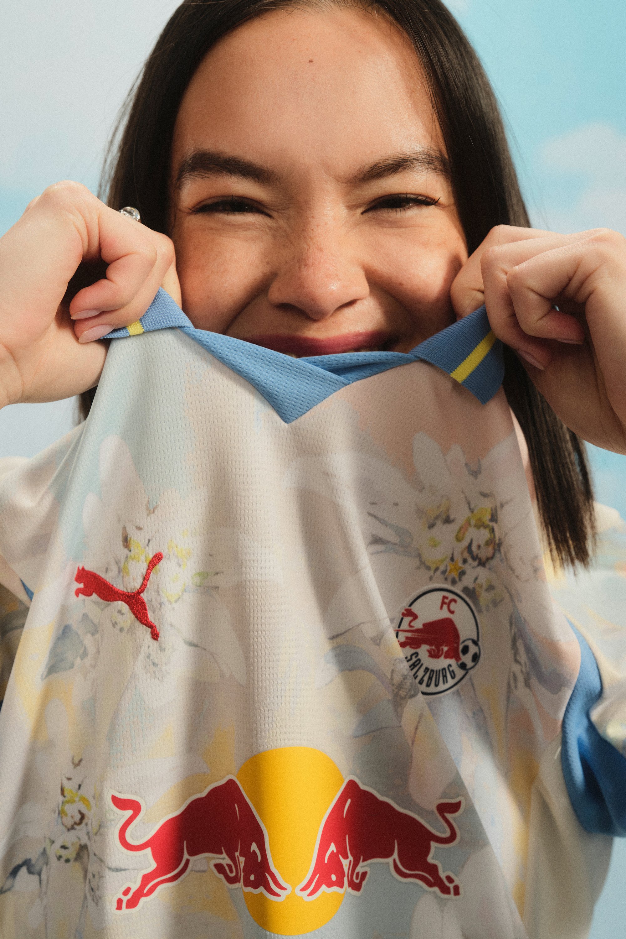 A young woman smiles while pulling a sports jersey towards her face, showcasing colorful team graphics and logos against a light background.