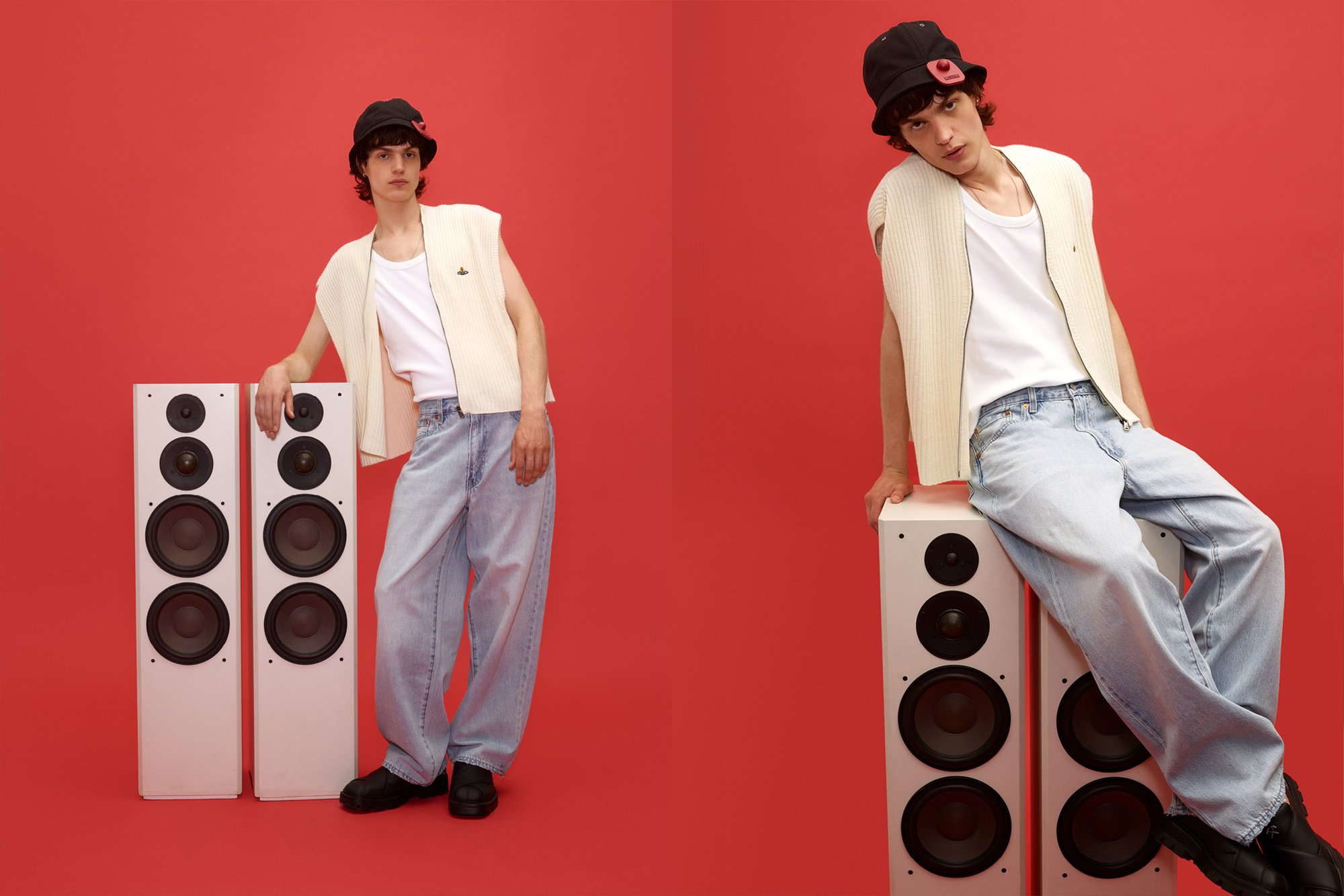 Joven con chaleco claro, camiseta blanca de tirantes, jeans holgados y gorra de cubo negra posa con dos altavoces blancos frente a un fondo rojo.