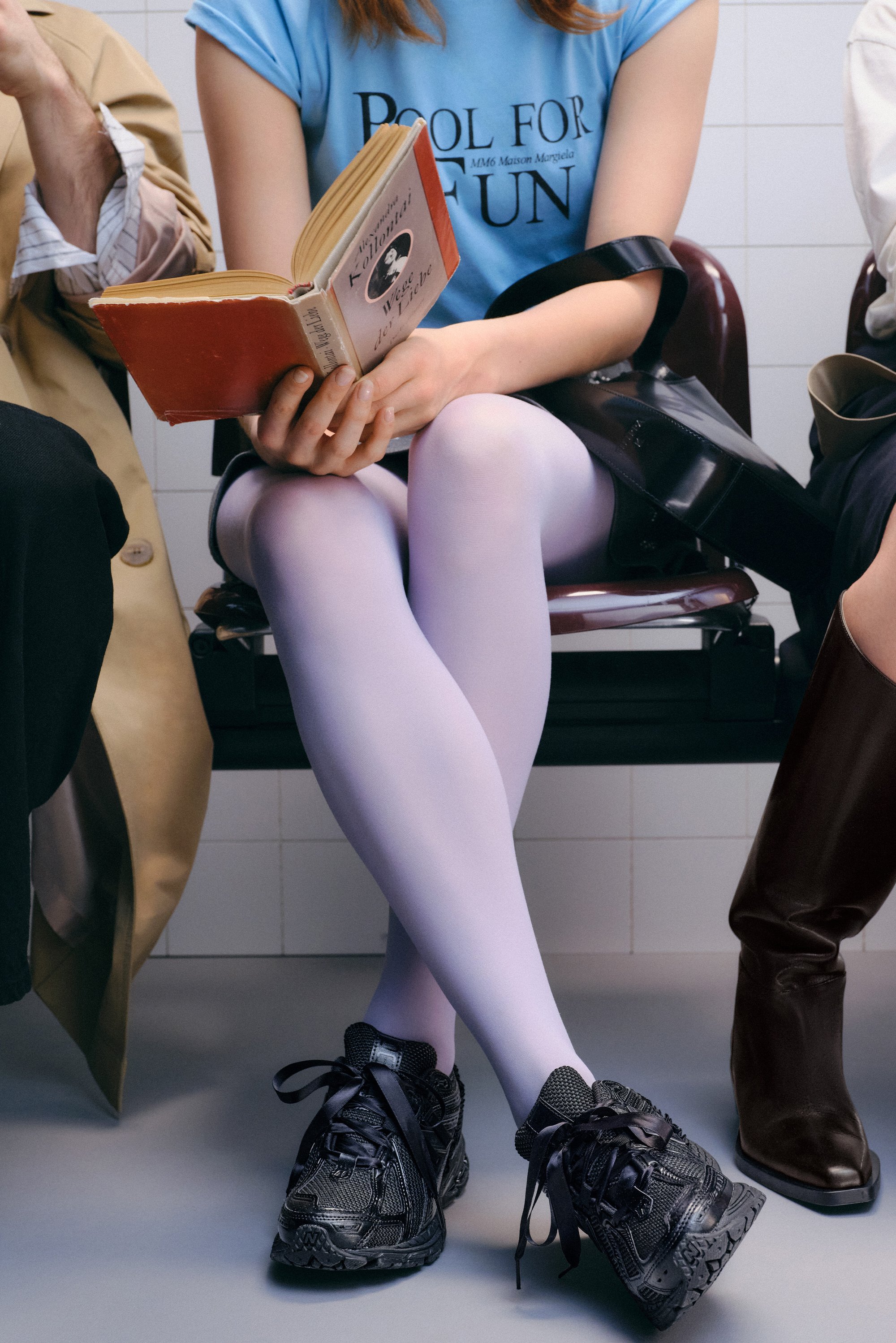 Una persona che indossa collant viola chiaro legge un libro mentre è seduta, affiancata da altre due persone in calzature e abbigliamento eleganti, in un ambiente con pavimento piastrellato.