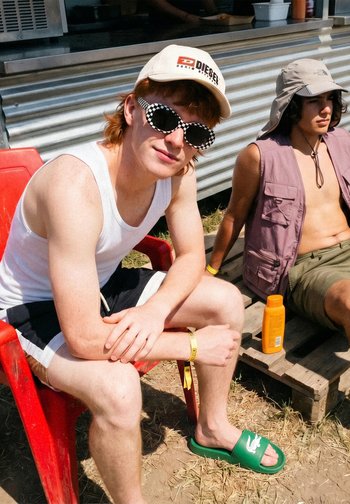 Zwei junge Männer sitzen im Freien; einer trägt ein weißes Tanktop, karierte Sonnenbrille und eine Kappe, der andere eine ärmellose Weste und einen Sonnenhut neben Sonnencreme.