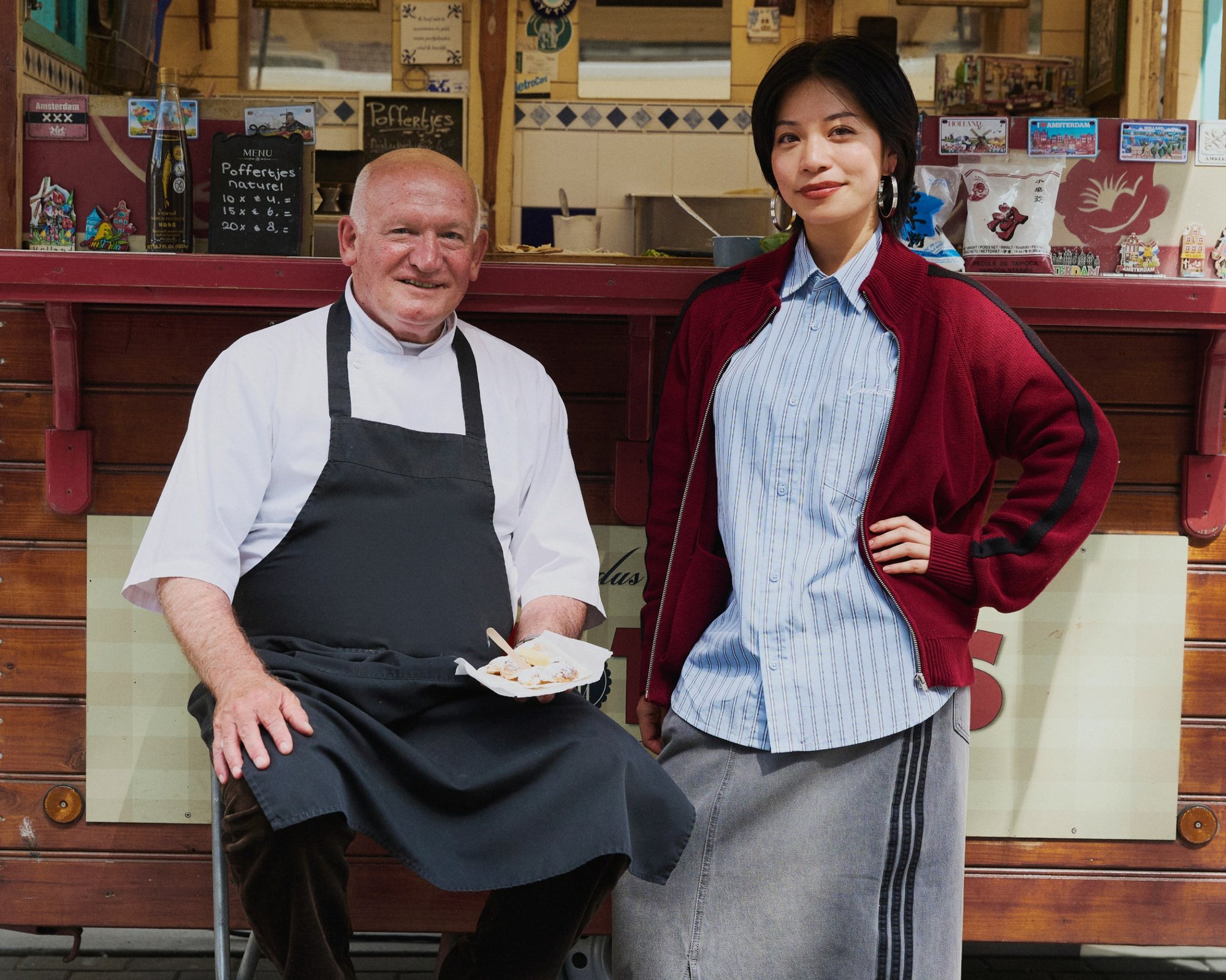 Deux personnes posent devant un stand de nourriture. L'homme porte une veste de chef blanche et un tablier noir, tenant un plat. La femme porte un cardigan rouge sur une chemise à rayures bleues et une jupe grise.