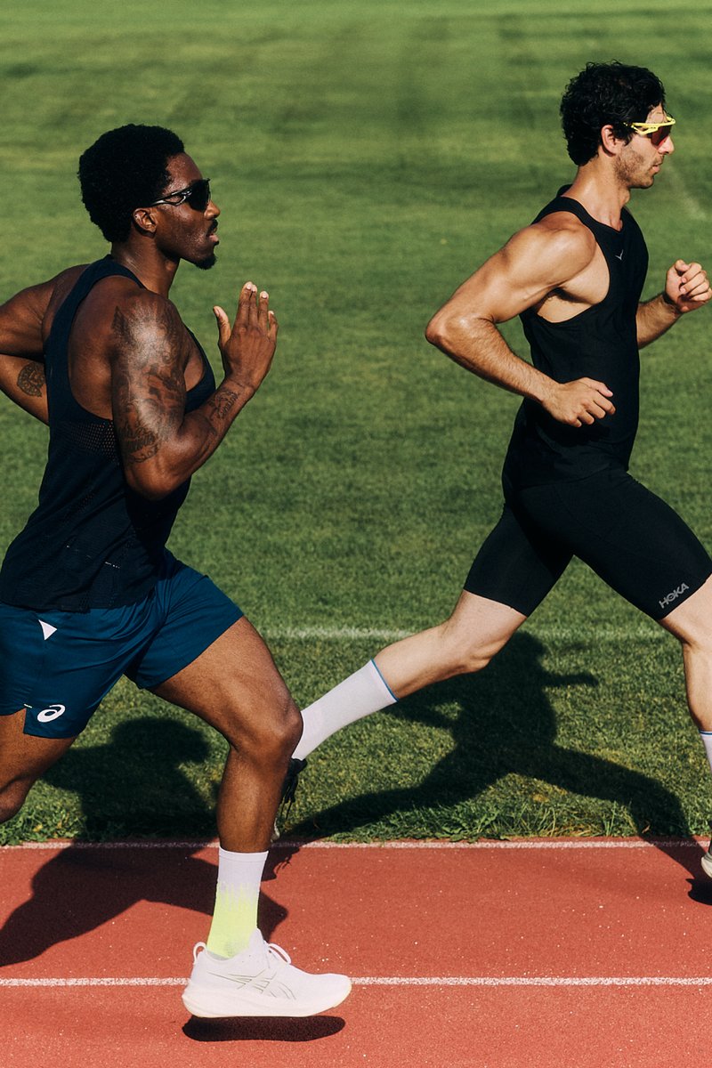 Three male runners in athletic gear sprint on a track, with green grass in the background. Sunlight enhances their focused expressions.