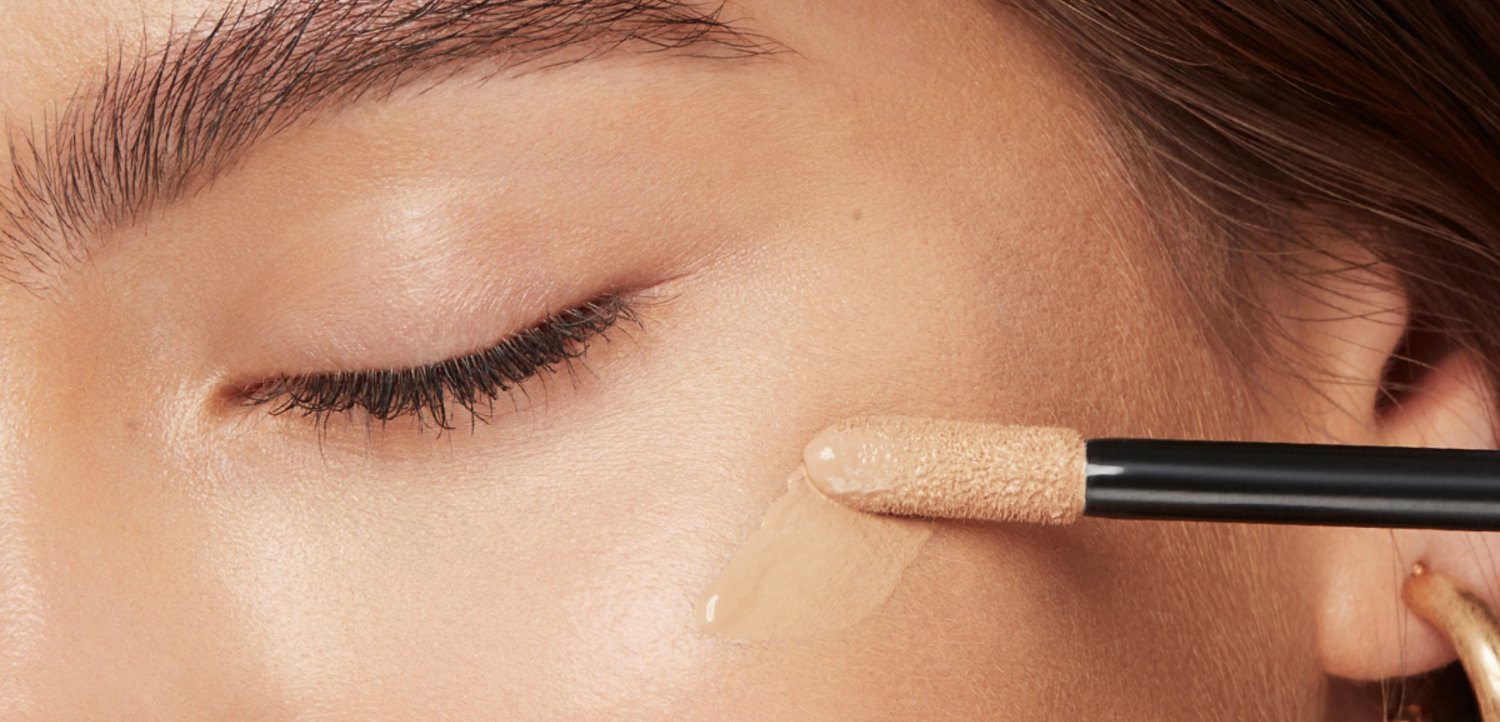 Œil fermé avec sourcil et cil visibles, applicateur de maquillage étalant un fond de teint liquide beige sur une peau lisse de la joue.