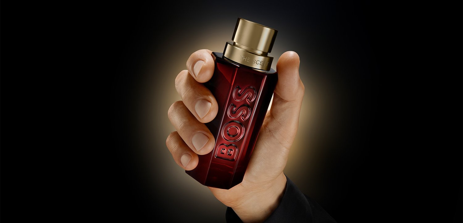 Mano che tiene una bottiglia rettangolare di profumo Boss di colore rosso scuro con tappo dorato su sfondo nero.