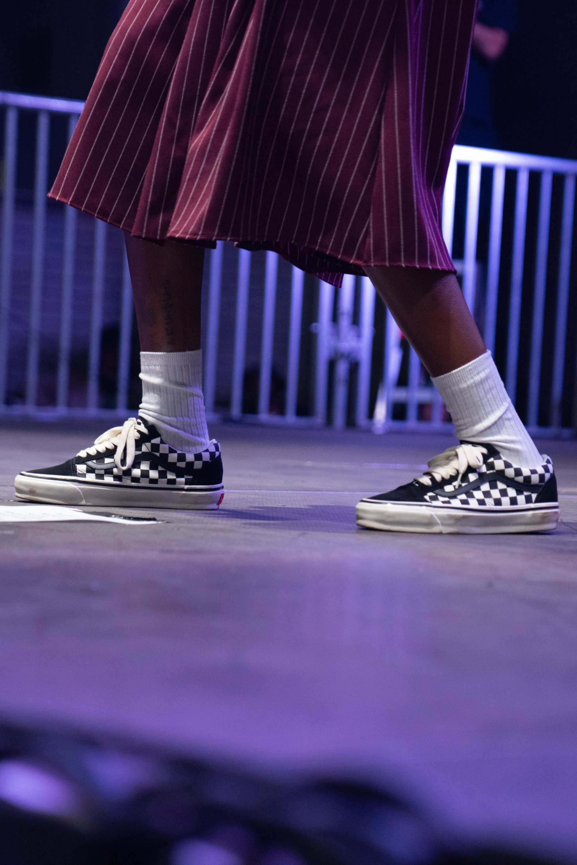 A person wearing a burgundy, striped skirt and checkered Vans sneakers stands on a stage with vibrant lighting.