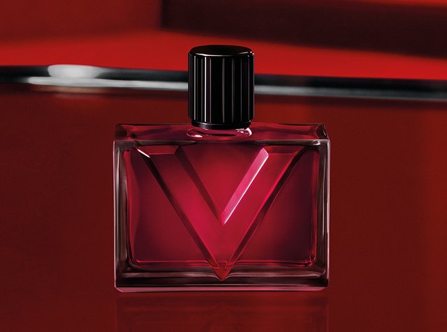 Flacon de parfum carré rouge avec un bouchon noir strié sur une surface et un fond rouge réfléchissants.