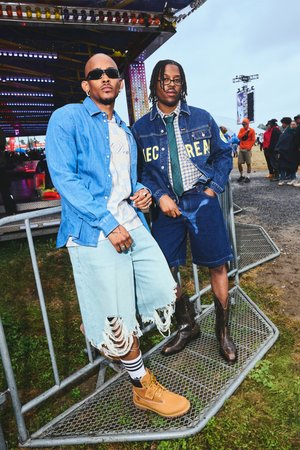 Zwei Männer posieren neben einem Metallzaun. Der eine trägt ein hellblaues Jeanshemd und zerrissene Shorts. Der andere hat eine Jeansjacke über einem karierten Hemd und dunklen Shorts.