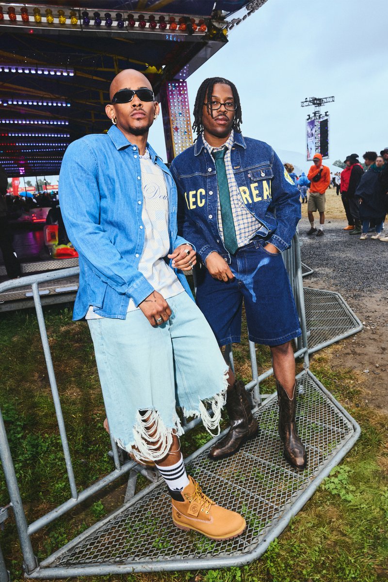 Zwei Männer posieren neben einem Metallzaun. Der eine trägt ein hellblaues Jeanshemd und zerrissene Shorts. Der andere hat eine Jeansjacke über einem karierten Hemd und dunklen Shorts.