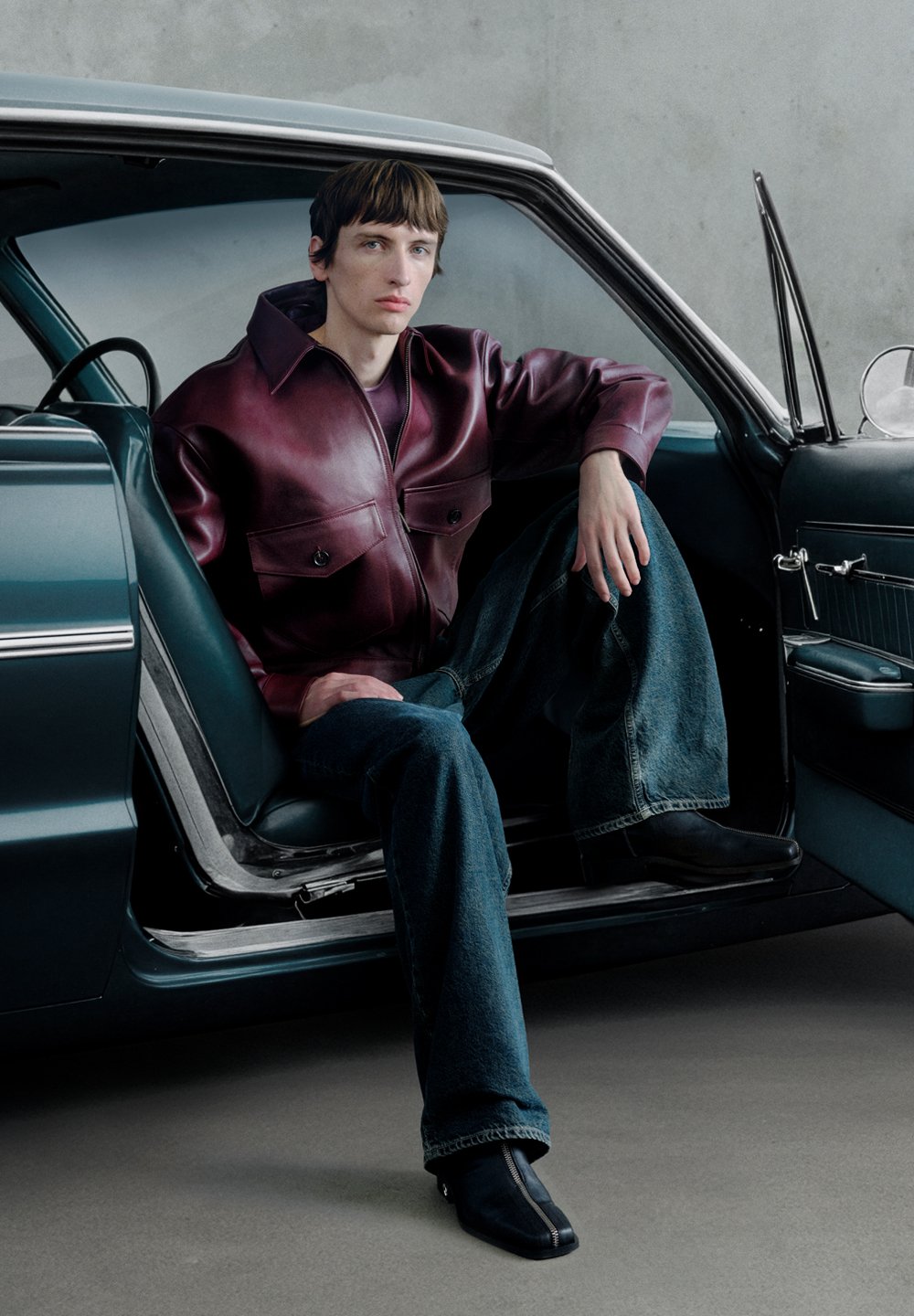 Casaco de cabedal cor castanho-avermelhado com dois bolsos, jeans de ganga azul e botas pretas até ao tornozelo. Posicionado no interior de um carro vintage.