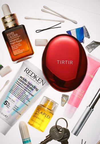 Collection of beauty products on a light background: glass serum bottle, red compact, cream tube, yellow oil, and keys, with cotton swabs nearby.