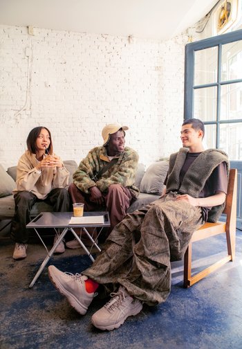 Three people share a casual conversation on a couch in a light-filled space, with drinks and a table between them.