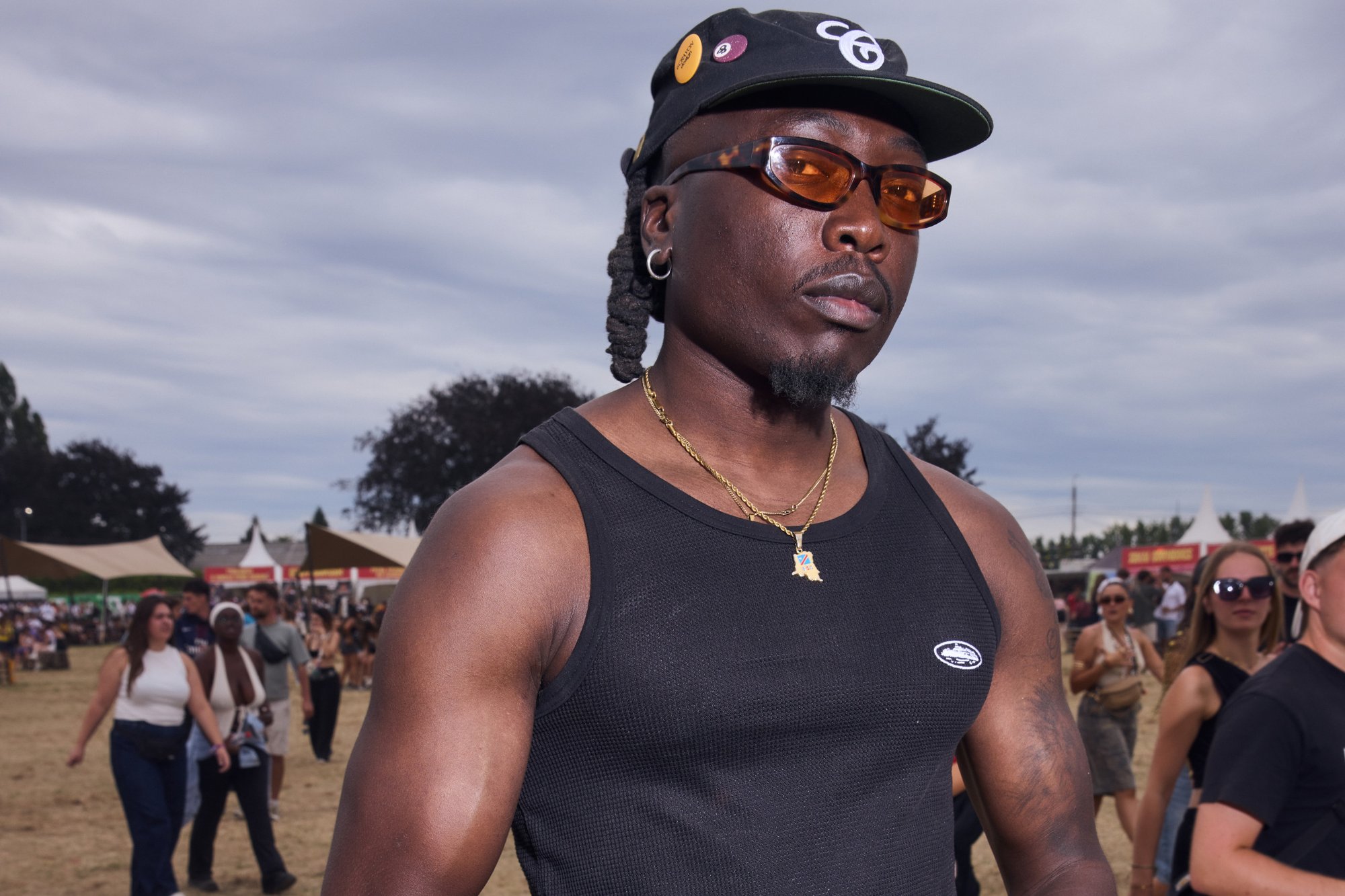 Débardeur noir, chaîne en or et pendentif ; casquette noire avec logo et épinglettes ; lunettes de soleil teintées orange ; peau texturée et cheveux tressés ; cadre de festival en plein air.