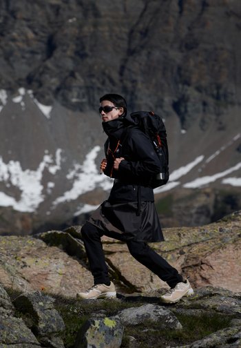 Eine Person mit Sonnenbrille und schwarzer Jacke wandert auf felsigem Terrain und trägt einen Rucksack, im Hintergrund sind schneebedeckte Berge.