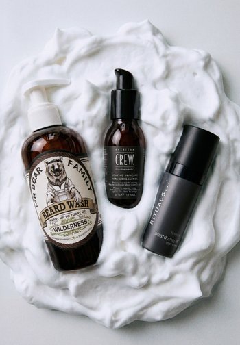 Tre skægplejeprodukter—skægvask, barberingsolie og skægformgiver—arrangeret på en hvid skummende barbercremebaggrund.