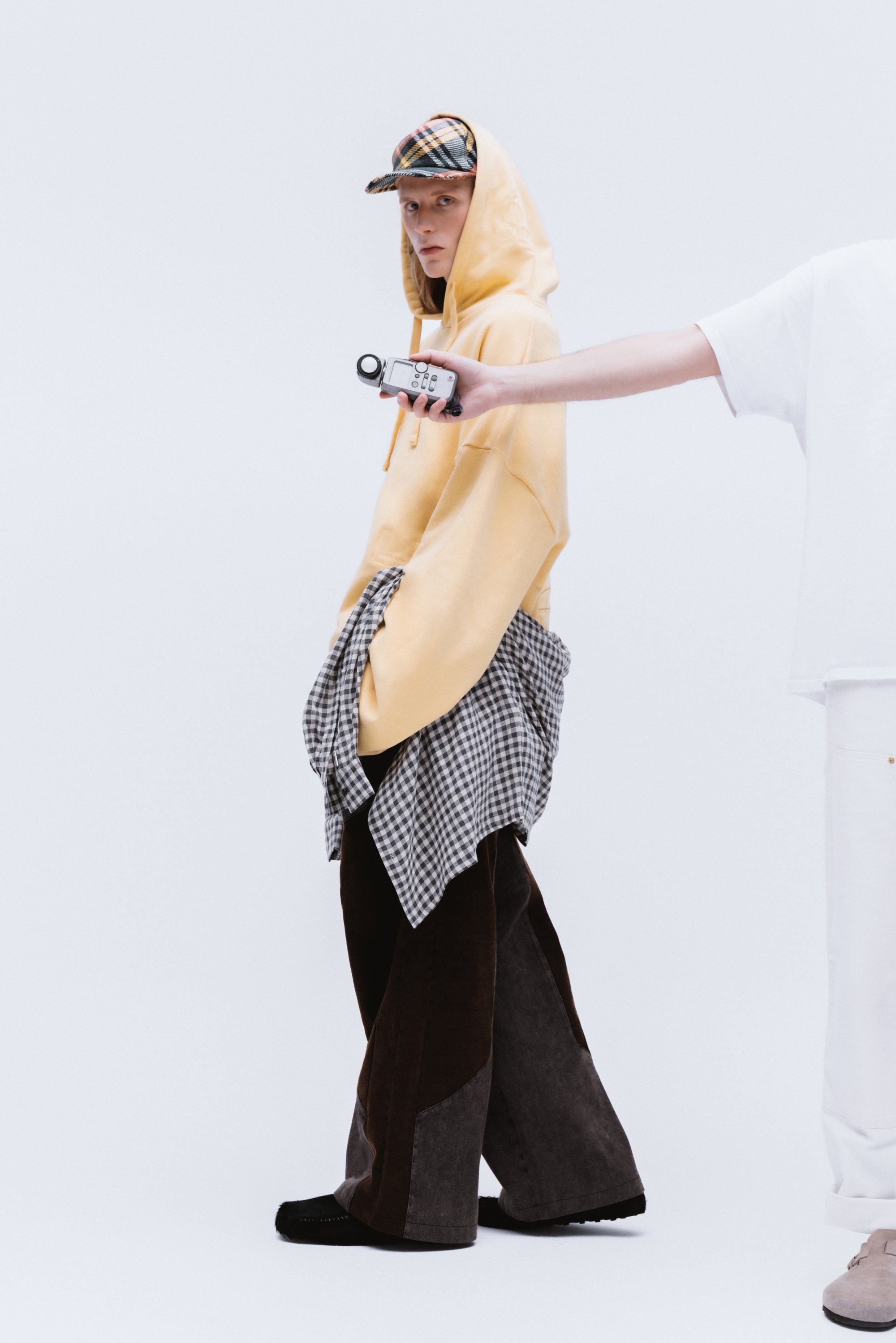 A model in a yellow hoodie and plaid cap poses, wearing layered oversized pants. Another person extends an object toward the camera.