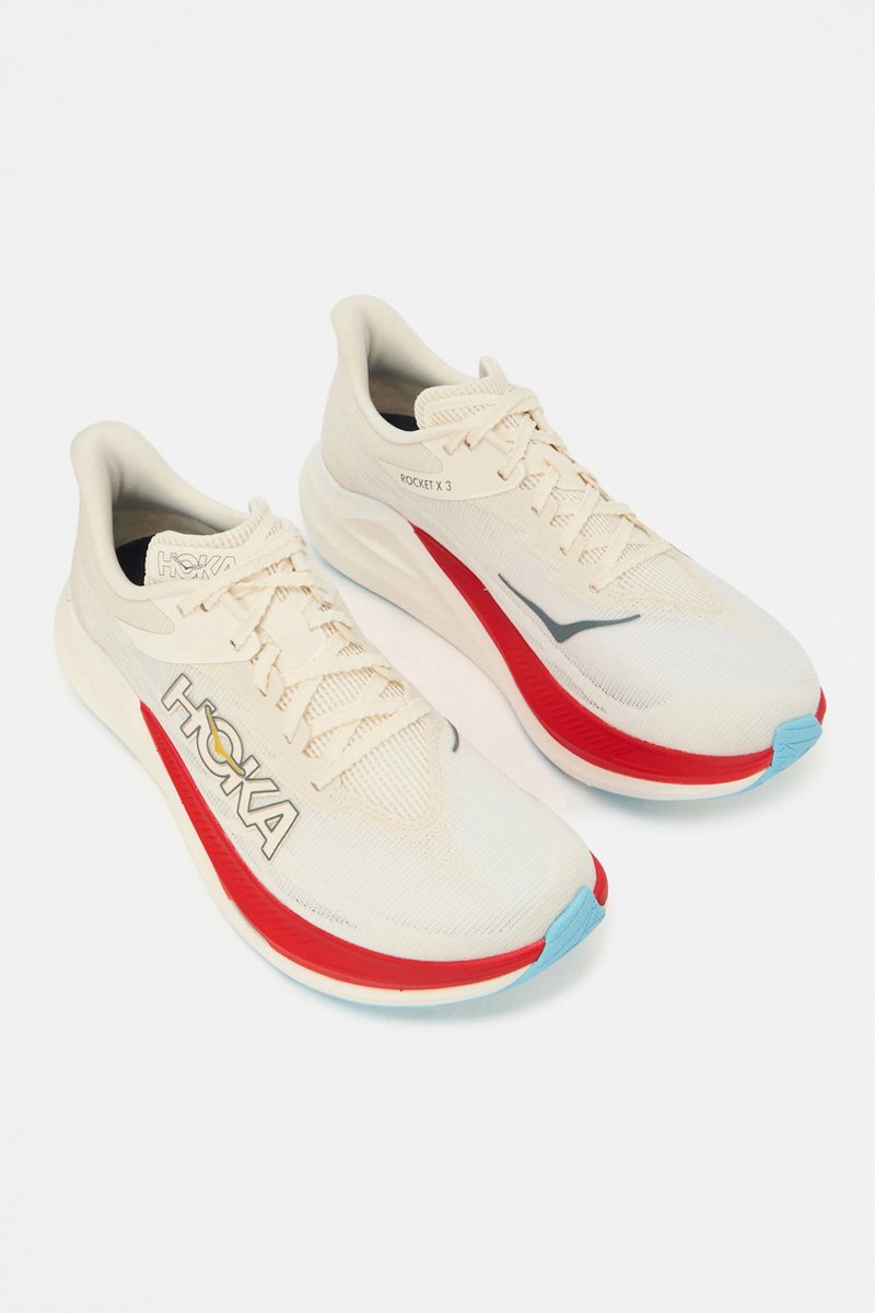 Pair of cream-coloured athletic running shoes with red and blue accents and "HOKA" logo on sides, on a plain white background.