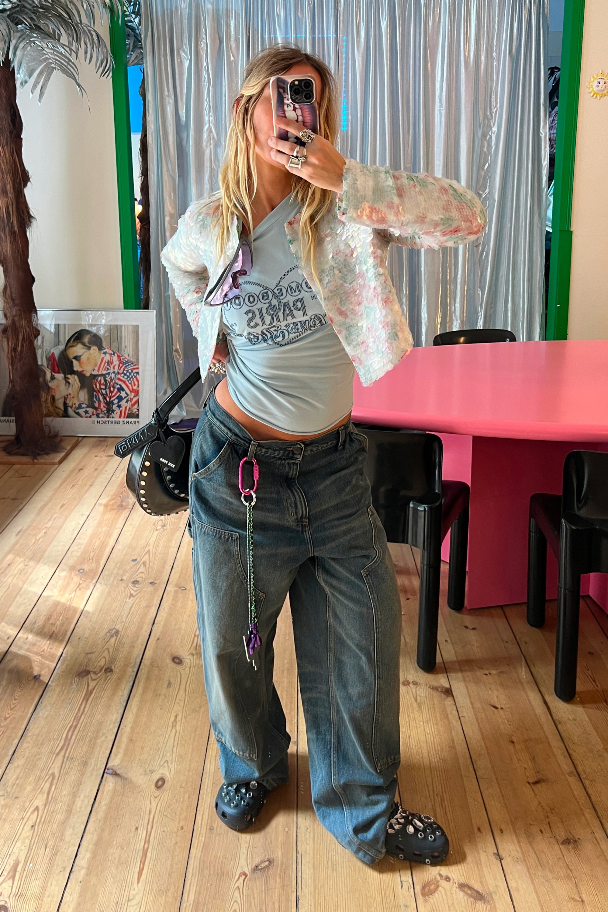 Persona con cabello largo y rubio que se está tomando un selfie en el espejo, vistiendo una chaqueta texturizada en tonos pastel, una camiseta gráfica azul, jeans holgados y zapatos negros con studs, en un ambiente interior.