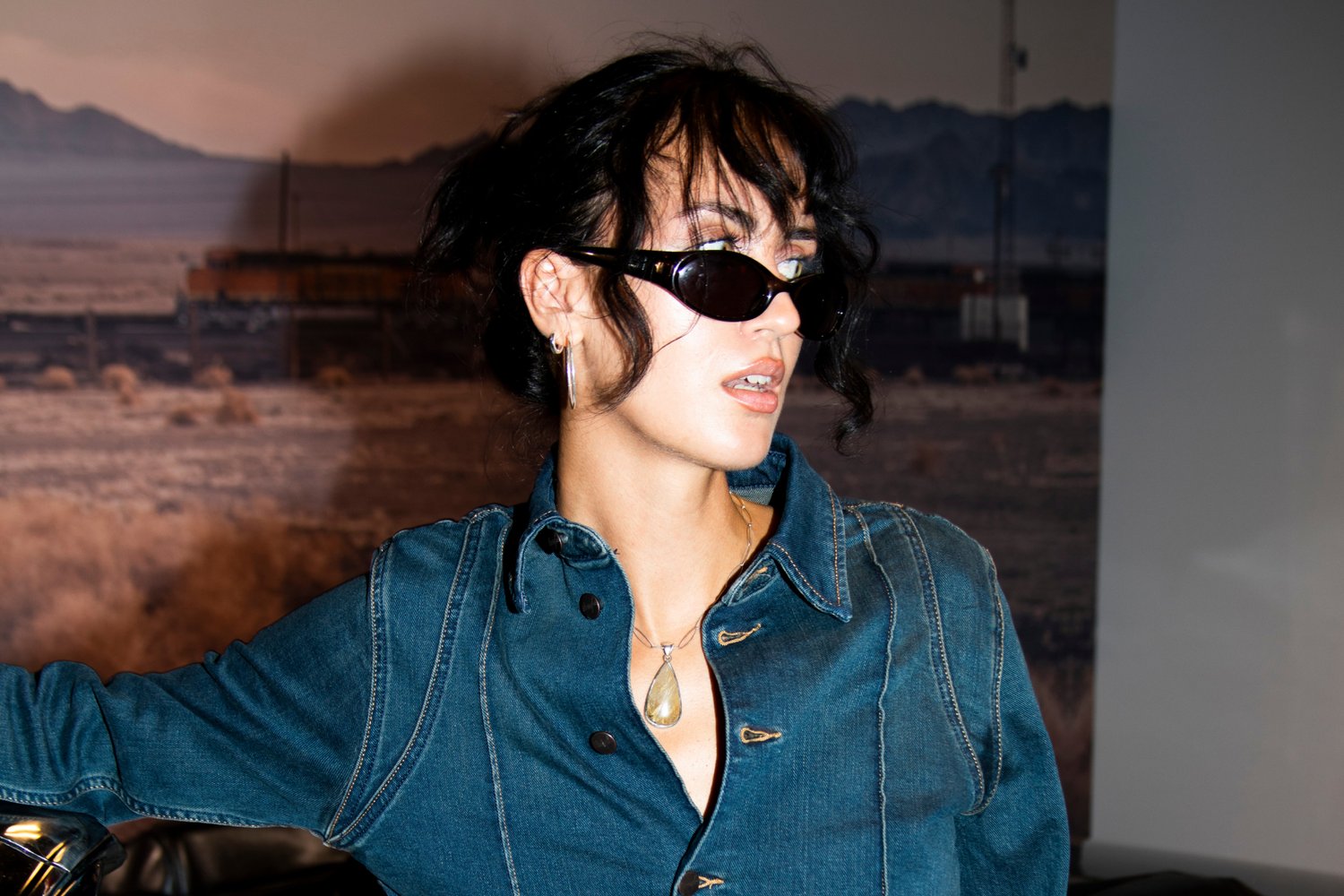 Zwarte zonnebril, spijkerjack met opvallende knopen en stiksels, gelaagde hangerketting, met golvend haar en een neutrale achtergrond.