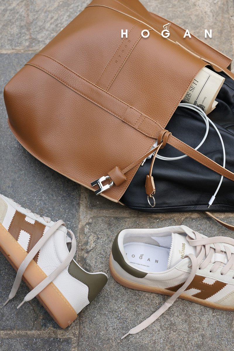 Hellbraune Lederhandtasche auf Steinboden mit schwarzem Beutel, Zeitung, weißen Ohrhörern und einem Paar weißen Sneakern mit braunen und grünen Akzenten.