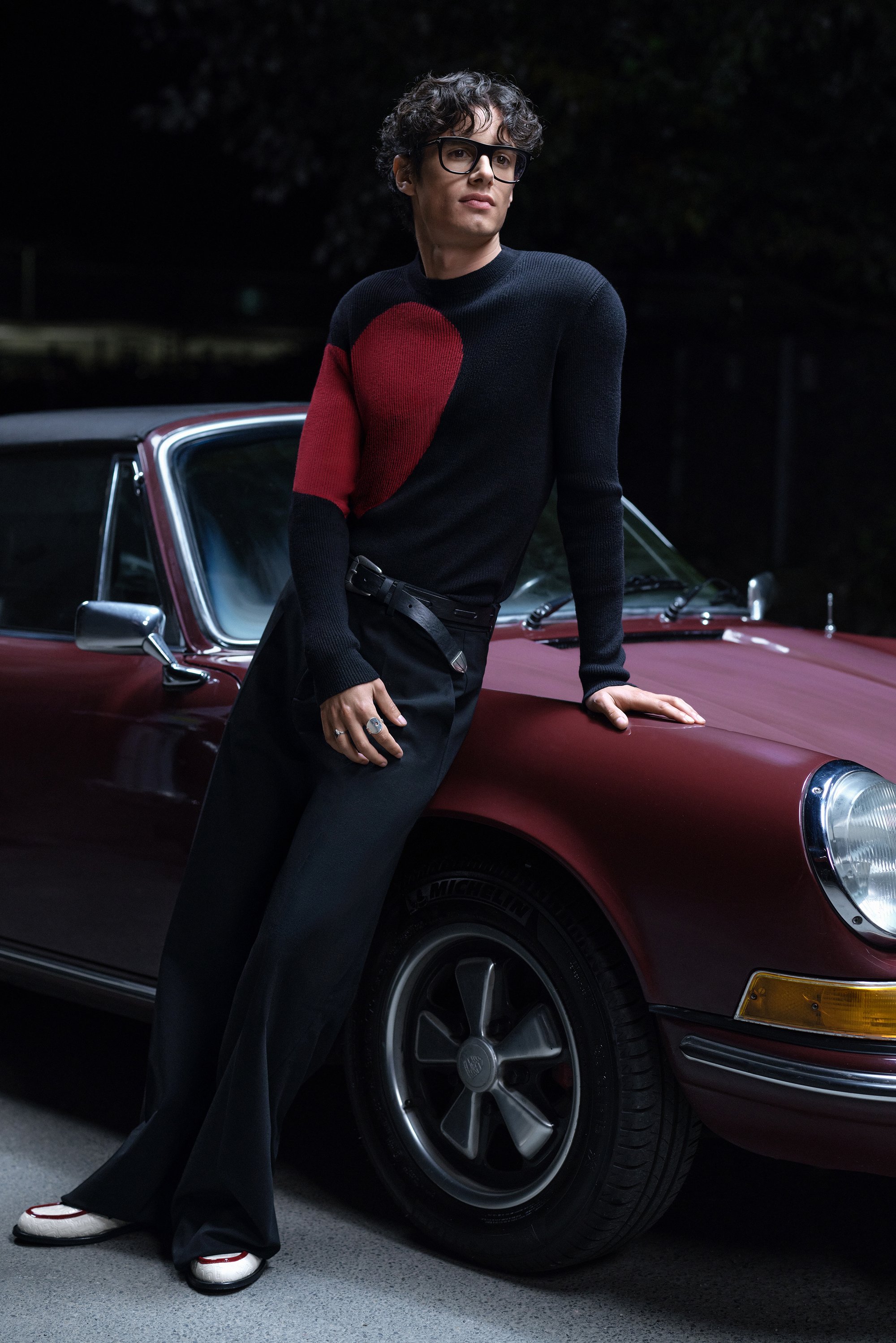 A young man with curly hair and glasses poses near a classic red car, wearing a black sweater with a red heart and black pants.