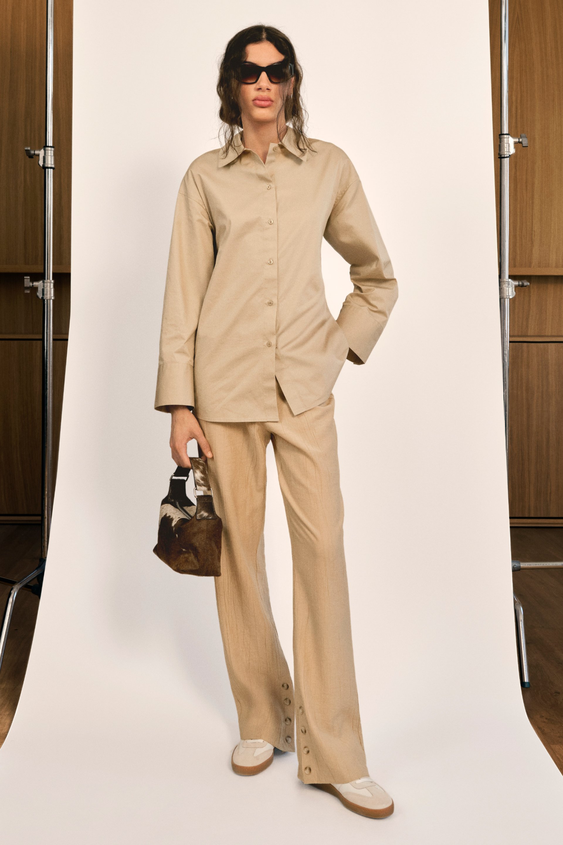 Una mujer se encuentra de pie contra un fondo neutro, vistiendo una camisa oversized beige y pantalones a juego, sosteniendo un pequeño bolso estampado.