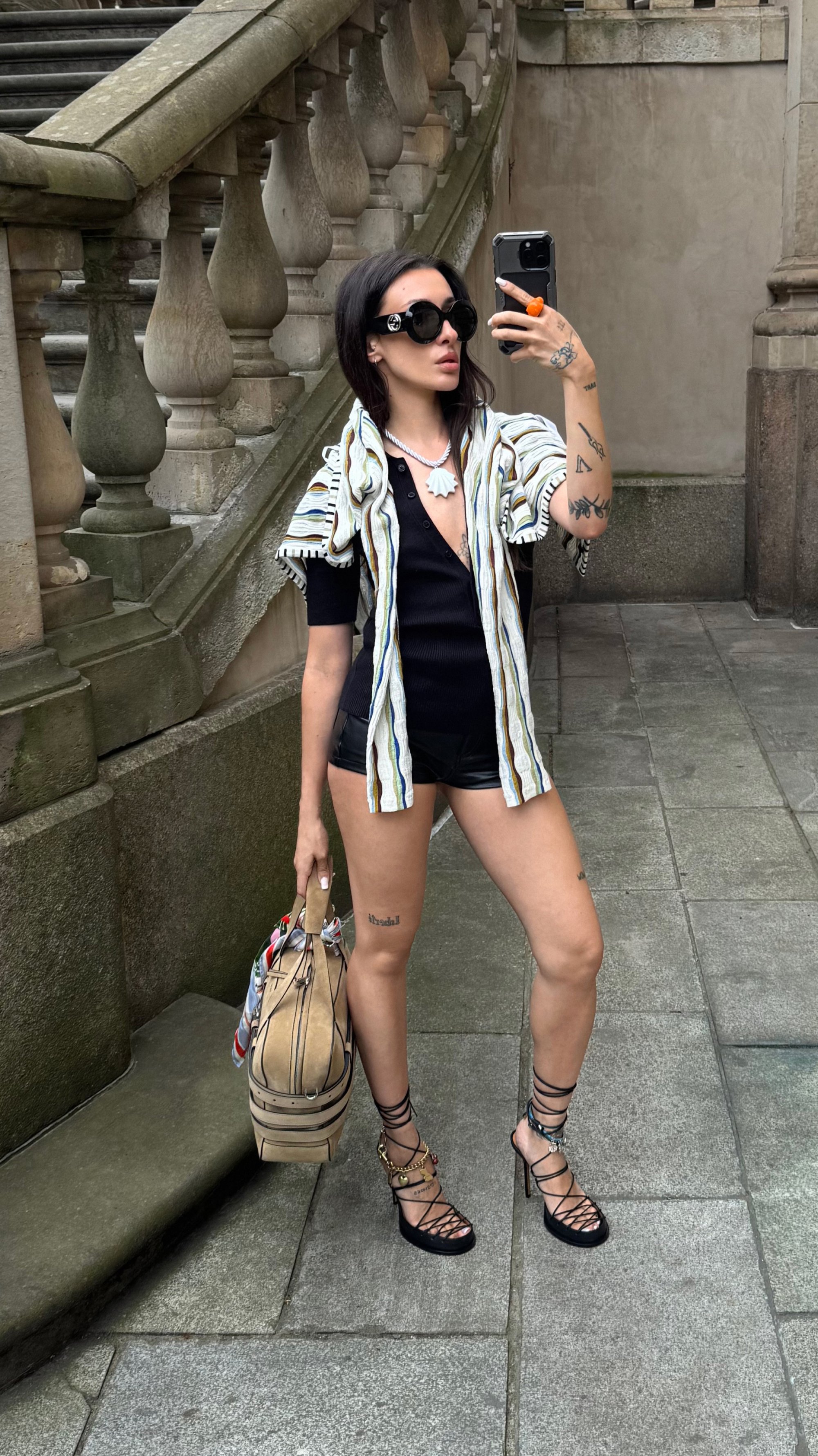 Borsa a mano tan con accenti a righe, top nero, camicia stampata, pantaloni in pelle e sandali con tacco alto e cinturini. Sfondo in pietra liscia.
