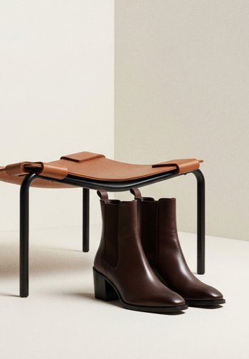 Brune skinnankelstøvler med blokkhæl og elastiske sidepaneler, plassert ved siden av en minimalistisk, brun skinn- og svart metallstol.