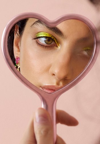 Gros plan sur le visage d'une femme avec un fard à paupières pailleté vert, des boucles d'oreilles en forme de fleur rose, reflété dans un miroir à main rose en forme de cœur.