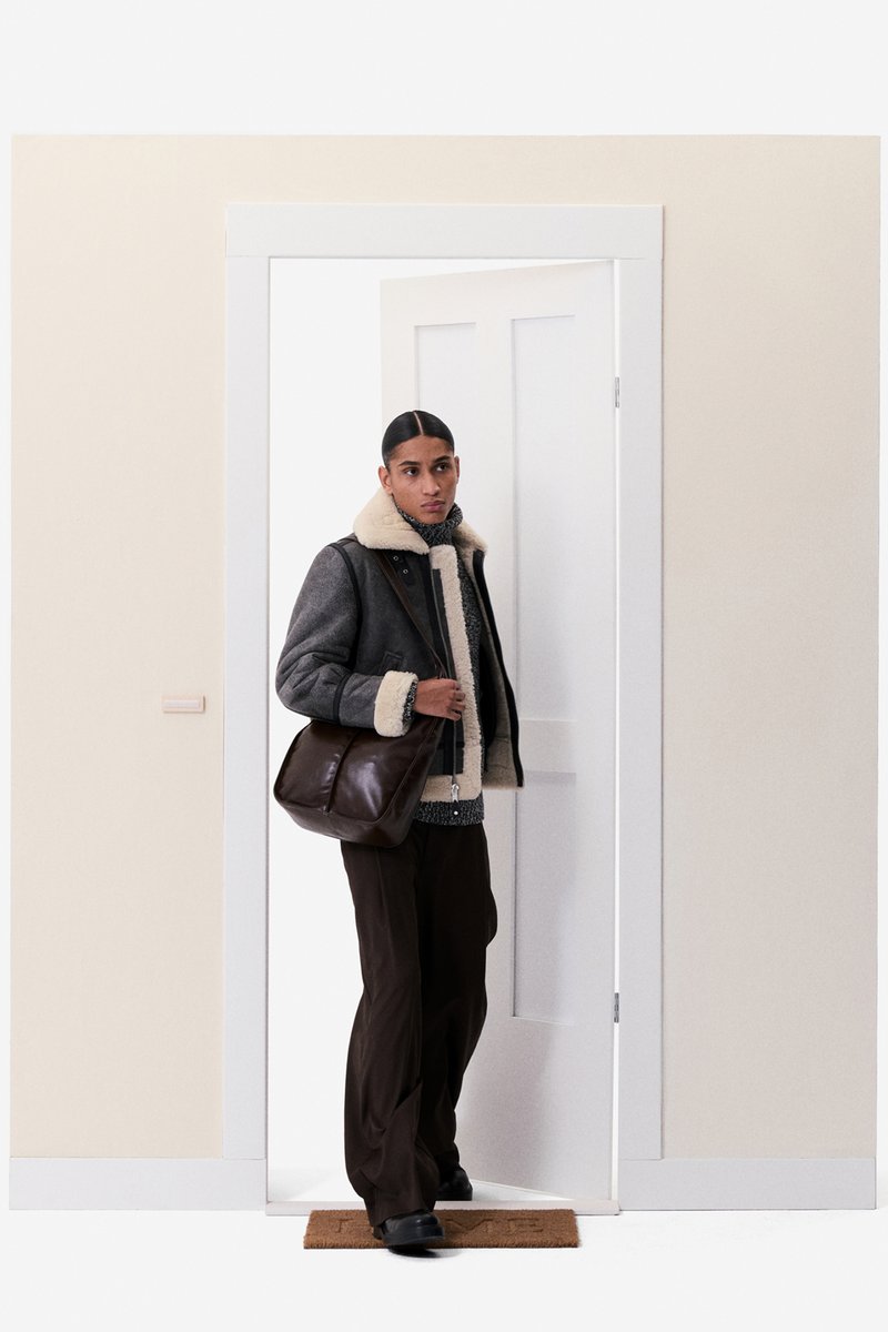 Gray shearling jacket with a cream collar, black and white patterned sweater, brown pants, and a brown leather messenger bag, standing by a door.