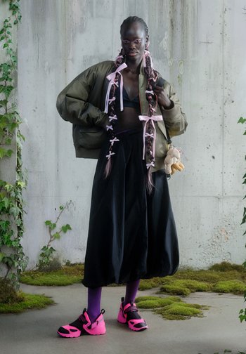 Πράσινο μπουφάν bomber, μαύρο κροπ τοπ, μαύρη φούστα· ροζ κορδέλες στα μαλλιά, ροζ και μαύρα παπούτσια, μωβ κάλτσες, αξεσουάρ αρκουδάκι.