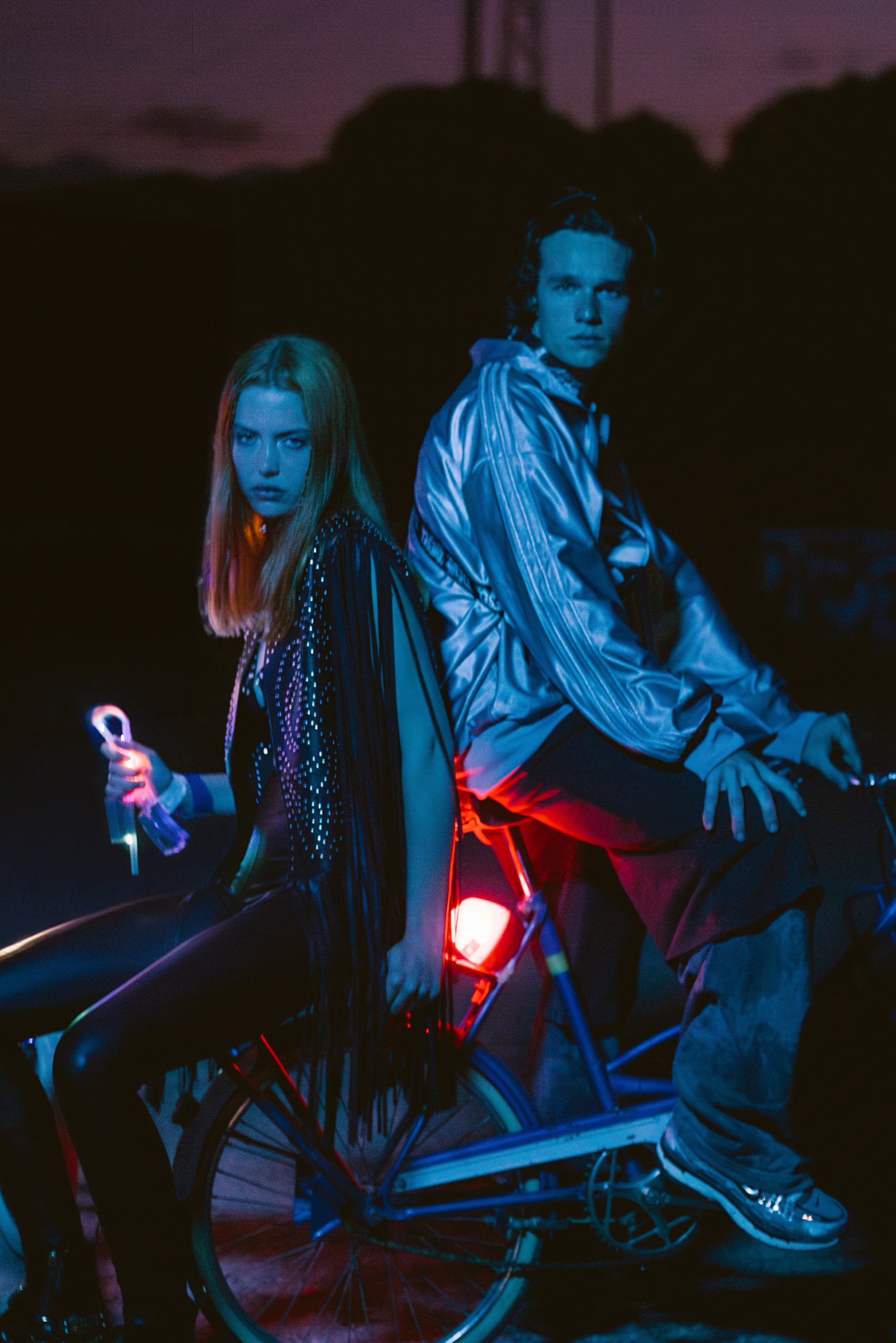 Twee jonge volwassenen zitten op een fiets bij schemerdonker, verlicht door blauwe en rode lichten. De vrouw houdt een gloeiend voorwerp vast en draagt een fringed vest.
