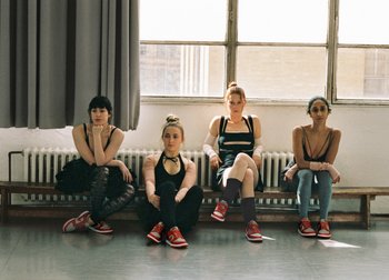 Four young women sit on a bench in a dance studio, wearing casual athletic outfits with red sneakers, against a backdrop of large windows.