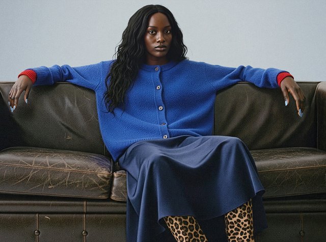 Blå ullkofta med röda detaljer, kombinerad med en blå kjol och leopardmönstrade stövlar. Vågigt svart hår, sittande på en mörk lädersoffa.