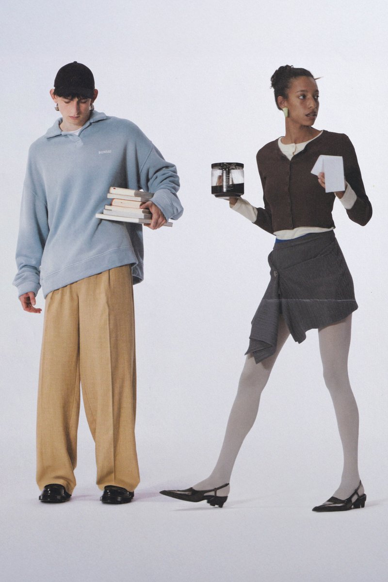 Gebreide donkere trui met een ritssluiting, lichtpaarse shirt en tan hoge taille broek. Eén persoon houdt papieren bekers vast, een ander schenkt uit een metalen kan.