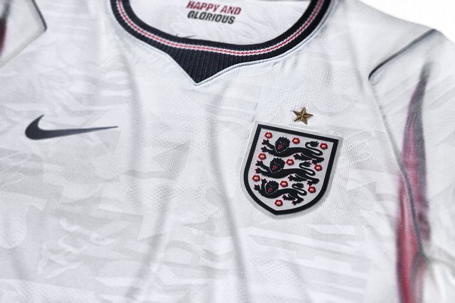 Biała koszulka piłkarska Anglii z herbem trzech lwów, jedną złotą gwiazdą, czarnym logo Nike oraz napisem "Happy and Glorious" wewnątrz kołnierzyka.