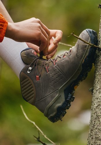 Hender som knyter skolissene på en brun tursko båret over hvite sokker, lent mot en trestamme med en pinne som sitter fast i sålen ute.